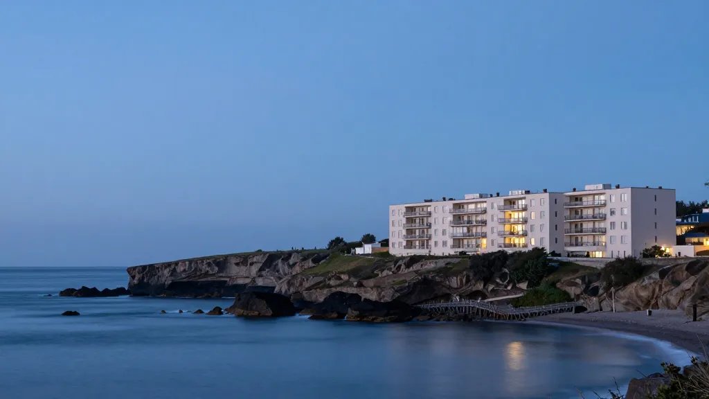 Wide shot of a single apartment building along a calm coastal cliffline at blue hour