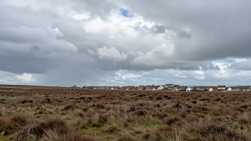 Expansive coastal moorland under dramatic skies, low-angle perspective over UK housing clusters and attic skylights in the distance