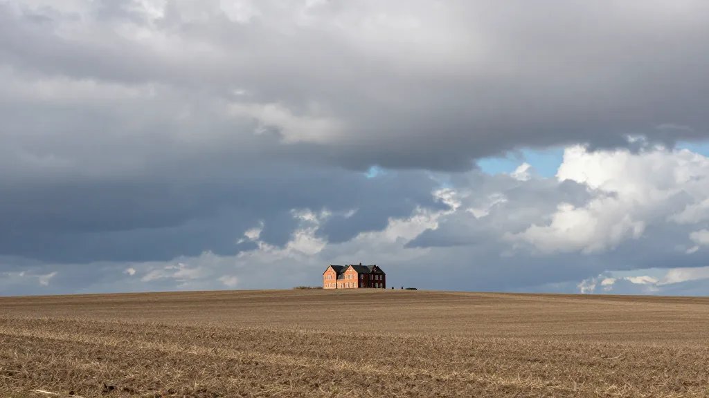 Vast rural field with a distant red-brick property silhouette under dramatic clouds