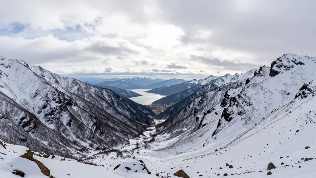 Snow-dusted alpine valley with distant lake under soft clouds